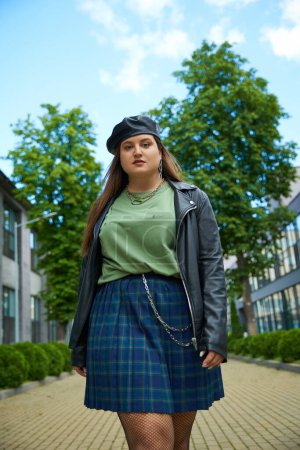 low angle view of confident woman with plus size body posing in leather jacket with black beret near blurred modern buildings and alley with trees on urban street outdoors, body positive 