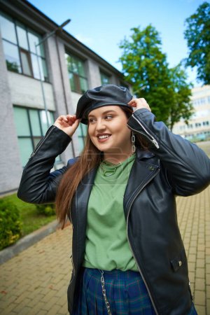 cheerful woman with plus size body posing in leather jacket and adjusting black beret near blurred modern building and trees on urban street outdoors, body positive, empowered and fashionable 