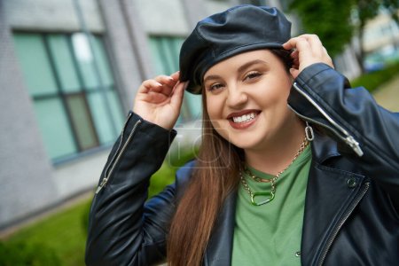 happy woman with plus size body posing in leather jacket and adjusting black beret near blurred modern building on urban street outdoors, body positive, urban chic, empowered and fashionable 