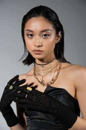 Porträt einer brünetten und asiatischen jungen Frau mit kurzen Haaren, die in schwarzem schulterfreien Kleid und Handschuhen mit goldenen Ringen posiert und auf grauen Hintergrund in die Kamera blickt, nasse Frisur, Halsketten, natürliches Make-up