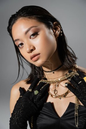 portrait de jeune femme brune et asiatique aux cheveux courts posant en gants noirs avec anneaux et robe bustier tout en tenant des bijoux dorés sur fond gris, coiffure mouillée, maquillage naturel
