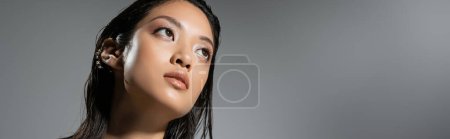 retrato de fascinante mujer joven asiática con el pelo corto y pendientes de oro posando mientras mira hacia otro lado sobre fondo gris, peinado húmedo, maquillaje natural, mirando hacia otro lado, bandera