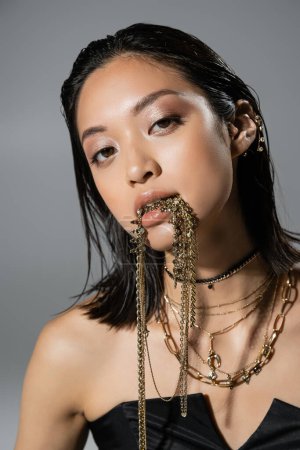 Foto de Retrato de la mujer joven asiática de moda con pelo corto posando en vestido negro sin tirantes mientras sostiene joyas de oro en la boca sobre fondo gris, peinado húmedo, maquillaje natural, mirando a la cámara - Imagen libre de derechos