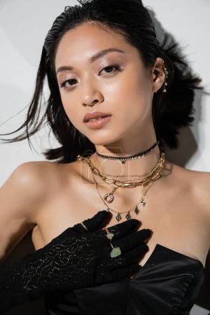 top view of alluring asian young woman with short hair lying in black gloves and strapless dress while posing in golden jewelry on grey background, wet hairstyle, natural makeup, looking at camera