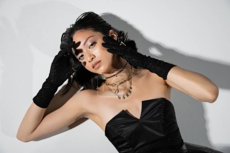 top view of mesmerizing young asian woman with short brunette hair lying in black gloves and strapless dress while posing in golden jewelry on grey background, wet hairstyle, hands near face 