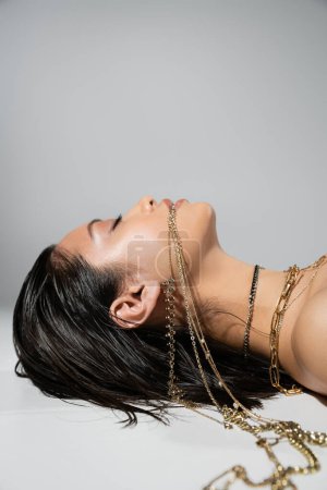 Foto de Vista lateral de mujer asiática joven con pelo corto morena sosteniendo joyas de oro en la boca mientras está acostado sobre fondo gris, maquillaje diario, peinado mojado, ojos cerrados - Imagen libre de derechos