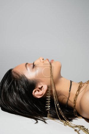 Téléchargez les photos : Jeune femme asiatique aux cheveux bruns courts tenant des bijoux dorés dans la bouche tout en posant sur fond gris, maquillage quotidien, coiffure humide, vue latérale, levant les yeux - en image libre de droit
