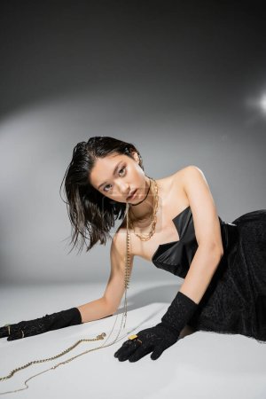 asian model with short brunette hair holding golden jewelry in mouth while looking at camera and posing in black strapless dress on grey background, everyday makeup, wet hairstyle, young woman 