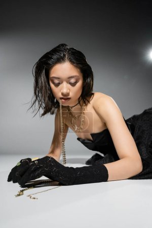 captivating asian model with short brunette hair holding golden jewelry in mouth while looking down and posing in strapless dress on grey background, everyday makeup, wet hairstyle, young woman 