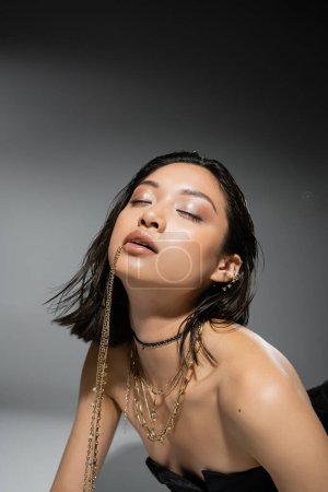 modèle asiatique élégant avec cheveux bruns courts tenant des bijoux dorés dans la bouche, posant en robe bustier sur fond gris, maquillage quotidien, coiffure humide, jeune femme, yeux fermés, portrait 