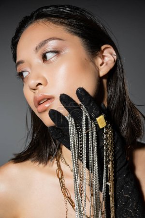 portrait of mesmerizing and asian young woman with short hair holding golden and silver jewelry while wearing glove and posing on grey background, wet hairstyle, natural makeup