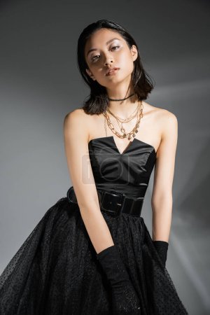 Porträt einer stilvollen asiatischen jungen Frau mit kurzen Haaren, die in einem schwarzen trägerlosen Kleid mit Gürtel und Handschuhen posiert, während sie in die Kamera auf grauem Hintergrund blickt, nasse Frisur, goldene Halsketten 