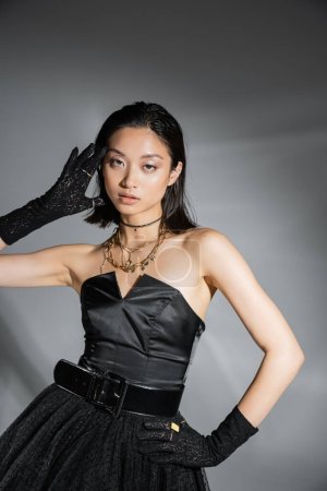 fascinante jeune femme asiatique aux cheveux courts posant avec la main sur la hanche en robe bustier noire avec ceinture et gants tout en regardant la caméra sur fond gris, coiffure humide, colliers dorés 