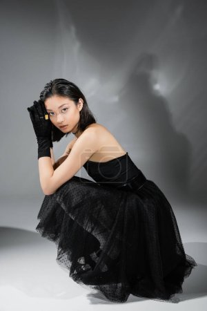 volle Länge hypnotisierende asiatische junge Frau mit kurzen Haaren sitzt in schwarzem trägerlosen Kleid mit Tüllrock und Handschuhen vor der Kamera auf grauem Hintergrund, nasse Frisur, goldene Halsketten 