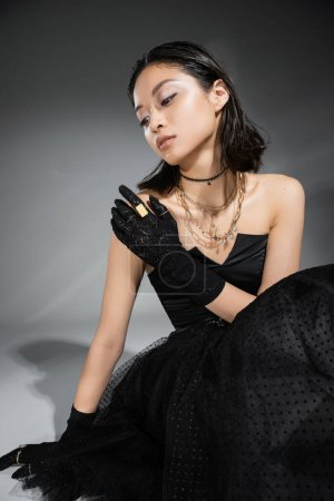 charming asian young woman with short hair sitting in black strapless dress with tulle skirt and gloves while looking away on grey background, wet hairstyle, golden necklaces and rings 