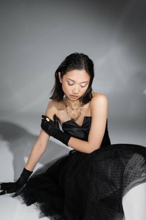 Foto de Impresionante mujer joven asiática con pelo corto sentado en vestido negro sin tirantes con falda de tul y guantes mientras mira hacia abajo sobre fondo gris, peinado mojado, collares y anillos de oro - Imagen libre de derechos