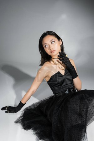 schicke asiatische junge Frau mit kurzen Haaren sitzt im schwarzen trägerlosen Kleid mit Tüllrock mit Gürtel und Handschuhen, während sie wegschaut und den Hals auf grauem Hintergrund berührt, nasse Frisur, goldene Halsketten 
