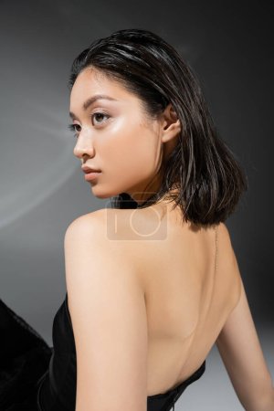 retrato de mujer joven asiática con pelo corto morena de pie en vestido negro sin tirantes sobre fondo gris, maquillaje diario, peinado mojado, modelo encantador, belleza natural 