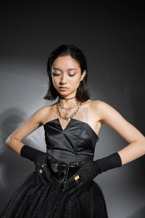 Foto de Encantadora mujer joven asiática con pelo corto posando en vestido negro sin tirantes con falda de tul mientras toca el cinturón y mirando hacia abajo sobre fondo gris, peinado mojado, joyas de oro - Imagen libre de derechos