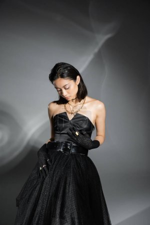 encantadora mujer joven asiática con pelo corto posando en vestido negro sin tirantes con falda de tul con cinturón y guantes de pie sobre fondo gris, peinado mojado, joyas de oro 