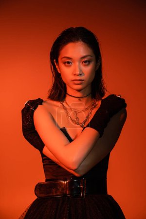 beautiful asian young woman with short hair and wet hairstyle posing with crossed arms in black strapless dress and gloves while standing on orange background with red lighting, golden jewelry 