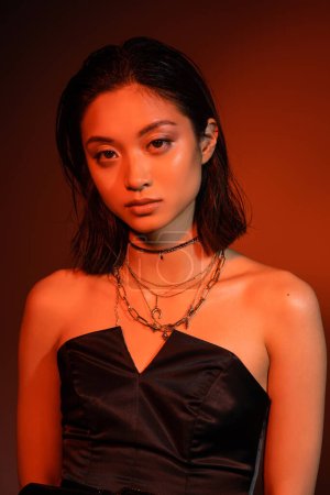 retrato de hermosa mujer joven asiática con pelo corto y peinado mojado posando en vestido negro sin tirantes mientras está de pie sobre fondo naranja con iluminación roja, joyas de oro 