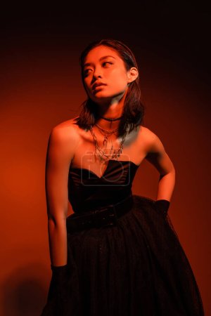 Photo for Asian woman with short hair and wet hairstyle posing with hand on hip in black strapless dress with tulle skirt and gloves while standing on orange background with red lighting, golden jewelry - Royalty Free Image