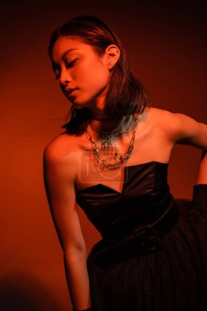 Photo for Mesmerizing asian woman with short hair and wet hairstyle posing in black strapless dress with tulle skirt and gloves while standing on orange background with red lighting, golden jewelry, young model - Royalty Free Image