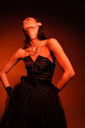 Foto de Mujer asiática elegante con los ojos cerrados y el peinado mojado posando en vestido negro sin tirantes con falda de tul y guantes, mientras que de pie sobre fondo naranja con iluminación roja, joyas de oro, modelo joven - Imagen libre de derechos
