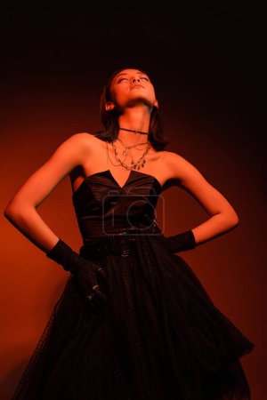 stylish asian woman with closed eyes and wet hairstyle posing with hands on hips in black strapless dress with tulle skirt and gloves while standing on orange background with red lighting, young model
