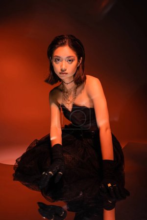 mujer asiática con pelo corto y peinado mojado posando en elegante vestido negro sin tirantes con falda de tul y guantes mientras está de pie sobre fondo naranja con iluminación roja, joyas de oro, modelo joven