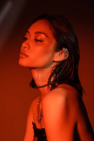 portrait of alluring asian woman with short hair and wet hairstyle posing in strapless dress with trendy cuff earring and necklaces on dark orange background with red lighting, young model 