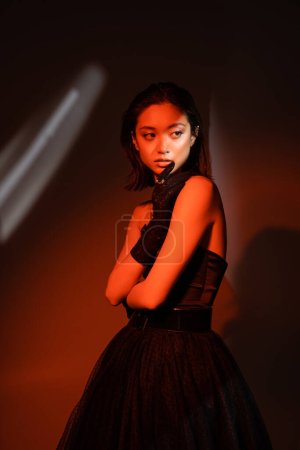 stunning asian woman with short hair and wet hairstyle posing in strapless dress and glove with trendy cuff earring and necklaces on dark orange background with red lighting, young model, looking away
