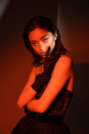 Photo for Portrait of stunning asian woman with short hair and wet hairstyle posing in strapless dress and glove with golden rings with trendy cuff earring on orange background with red lighting, young model - Royalty Free Image