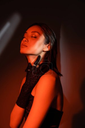 Photo for Portrait of beautiful asian model with closed eyes and wet hairstyle posing in strapless dress and glove with golden rings and touching neck on dark orange background with red lighting, young woman - Royalty Free Image