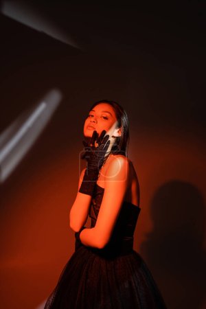 attractive asian woman with wet hairstyle posing in strapless dress with tulle skirt and black gloves with rings while standing on dark orange background with red lighting, model, looking at camera 