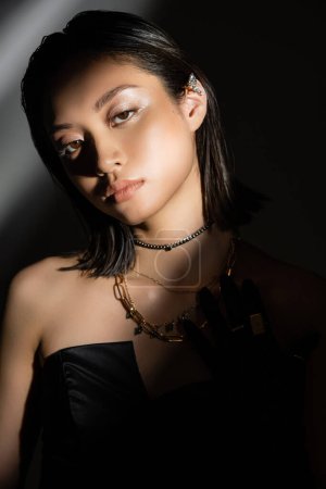 Porträt einer bezaubernden Asiatin mit feuchter Frisur und kurzen Haaren, die im schulterfreien Kleid posiert, während sie auf grauem Hintergrund steht, junges Model, in die Kamera blickend, Schatten, Dunkelheit