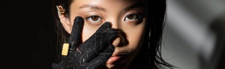 Téléchargez les photos : Portrait de jeune femme asiatique avec coiffure mouillée et cheveux courts en gant noir avec anneaux dorés touchant le visage tout en se tenant sur fond gris, modèle, regardant caméra, ombres, sombre, bannière - en image libre de droit
