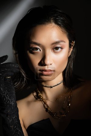 portrait of enchanting asian young woman with wet hairstyle and short hair posing in black glove while standing on grey background, model, looking at camera, shadows, dark, shimmer eyeshadow