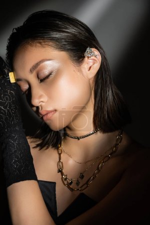 portrait de jeune femme asiatique enchanteresse à la coiffure mouillée et aux cheveux courts posant en gant noir avec anneau et manchette d'oreille debout sur fond gris, jeune mannequin, yeux fermés, ombres, sombre