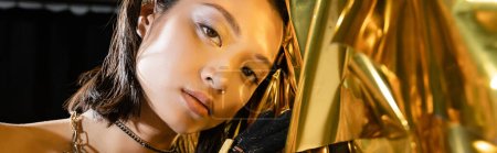 Foto de Retrato de seductora mujer joven asiática con el pelo corto mojado y joyas posando junto a fondo dorado brillante, modelo, mirando a la cámara, lámina arrugada, belleza asiática natural, pancarta - Imagen libre de derechos