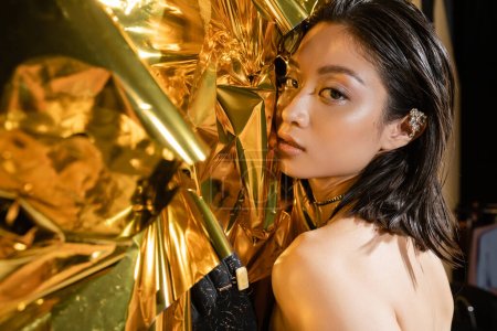 portrait de séduisante jeune femme asiatique aux cheveux courts mouillés posant à côté d'un fond doré brillant, mannequin, regardant la caméra, feuille jaune ridée, beauté naturelle, gant noir 