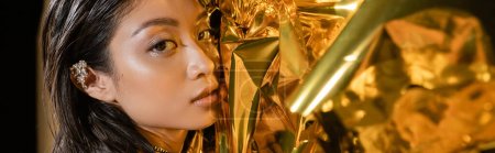 Porträt einer verführerischen asiatischen jungen Frau mit nassen kurzen Haaren und Ohrenmanschette, die vor leuchtend gelbem Hintergrund posiert, Modell, in die Kamera blickend, faltige Goldfolie, natürliche Schönheit, Banner 