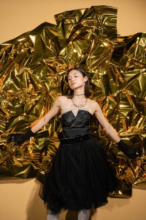 bezaubernde asiatische junge Frau mit kurzen Haaren posiert in schwarzem trägerlosen Kleid mit Tüllrock und Handschuhen, während sie neben leuchtend gelbem Hintergrund steht, Modell, faltige goldene Folie, schaut weg