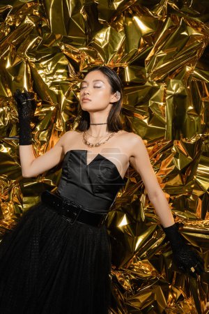 attractive asian young woman with short hair and closed eyes posing in black strapless dress with tulle skirt and gloves while standing next to shiny golden background, model, wrinkled foil