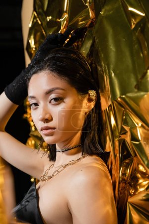 elegante asiatische junge Frau mit nasser Frisur und kurzen Haaren posiert im schulterfreien Kleid mit schwarzem Handschuh neben goldenem Hintergrund, Model, blickt in die Kamera, faltige gelbe Folie