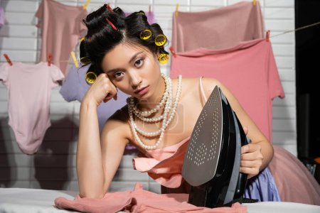 tired asian housewife with hair curlers in pink ruffled top and pearl necklace looking at camera while holding iron near clean clothes hanging on blurred background, housework, young woman, laundry 