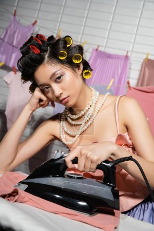 müde asiatische Hausfrau mit Lockenwicklern in rosa Rüschen Top und Perlenkette Bügeln und Blick in die Kamera in der Nähe nasse Kleidung hängen auf verschwommenem Hintergrund, Hausarbeit, junge Frau, Wäsche 