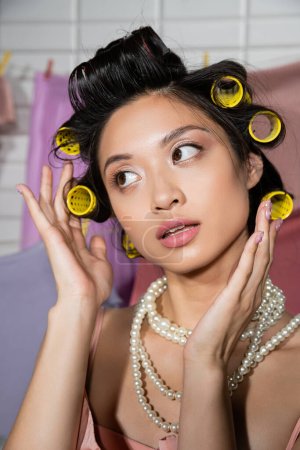 zamyślona młoda azjatycka kobieta dotykająca włosów lokówkami do włosów i pozująca w perłowym naszyjniku w pobliżu czyste i mokre pranie wiszące na rozmytym tle, prace domowe, gospodyni domowa, odwracając wzrok