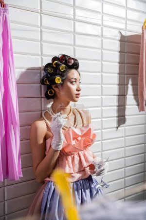 Téléchargez les photos : Asiatique jeune femme avec des bigoudis de cheveux debout en haut à volants rose, collier de perles et gants fumant et tenant le verre près de buanderie humide suspendue près de tuiles blanches, fer, cigarette, femme au foyer - en image libre de droit